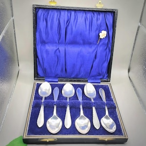 May include: A set of six silver spoons presented in a black box with a blue velvet lining. The spoons feature a traditional design with rounded bowls and elegant handles. A small white flower pin is attached inside the box.