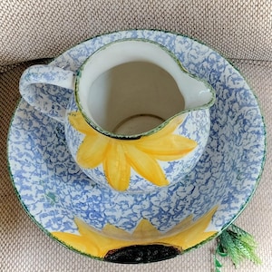 May include: A ceramic pitcher and matching basin set featuring a blue and white sponge-painted pattern. Both pieces are decorated with large, hand-painted yellow sunflower motifs with dark brown centers. The pitcher has a curved handle and spout.
