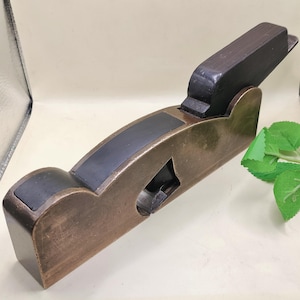 May include: A vintage bronze-toned hand plane with a dark wooden handle and accents. The plane features a curved body with a black insert and a blade opening. The tool is designed for woodworking and shaping materials.