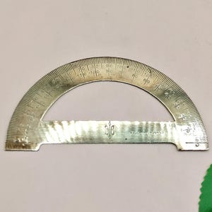 May include: A silver-colored protractor with degree markings from 0 to 180. The tool is semi-circular with a flat base and has the text "MADE IN ENGLAND". The protractor is used for measuring angles.