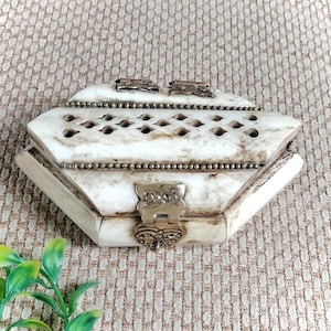 May include: A hexagonal box crafted from off-white, marbled material featuring a lattice-cut lid, metallic beaded trim, and a central silver-toned metal latch. The box has two small metal hinges at the back and a smooth, polished surface finish.