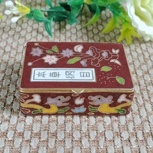 以下が含まれることがあります： 長方形の小箱。マルーン色で、金色の縁取りと蝶番付きの蓋が付いています。蓋には花と葉のデザイン、白い長方形の中に漢字が描かれています。箱の側面は龍のモチーフで装飾されています。