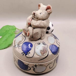 May include: A ceramic trinket box with a lid, featuring a light grey teddy bear figurine sitting on top. The box is decorated with blue and white balloon designs. A small black and white football is next to the bear.