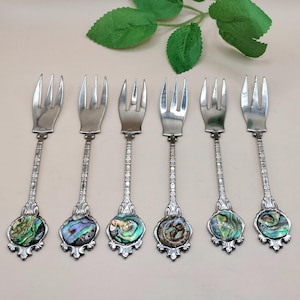 May include: Six silver forks with ornate handles and abalone shell accents. The forks have three prongs and decorative stems. The abalone shells display iridescent colours of green, blue, and purple. The forks are arranged in a row.