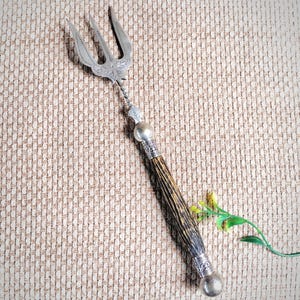 May include: A three-pronged silver serving fork featuring an ornate engraved metal head, a twisted metal neck, and a textured dark brown handle with silver-toned metal caps at both ends, finished with a rounded silver-toned metal pommel.