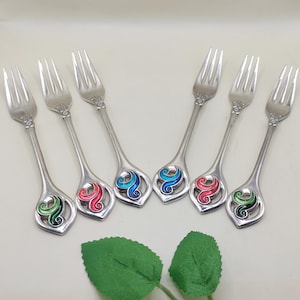 May include: Set of six silver forks with decorative handles. Each handle features a colorful, swirling design in green, red, and blue. The forks are arranged in a fan shape, showcasing their elegant design.
