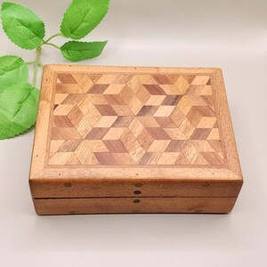 Peut inclure: Une boîte en bois rectangulaire avec un motif géométrique sur le couvercle. Le couvercle présente une mosaïque de pièces de bois claires et foncées disposées en un motif de cube tridimensionnel répétitif. La boîte est en bois aux tons chauds.