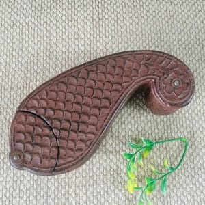 May include: A brown, fish-shaped wooden box with intricate carved details. The box features a scale pattern on the main body and floral carvings on the curved end. A small metal hinge and clasp secure the lid. The box is displayed on a textured, neutral-toned surface.