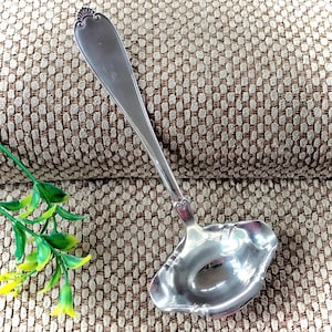 May include: A silver serving ladle with a decorative handle and a scalloped bowl. The ladle is made of a shiny metal and is designed for serving soups or sauces. The handle has an ornate design at the top.
