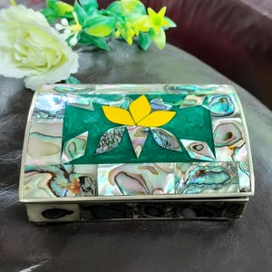 Peut inclure: Une boîte à bijoux rectangulaire avec un cadre argenté et un couvercle décoré de morceaux de coquille d'ormeau irisés. Le centre présente une incrustation verte avec un motif de fleur jaune. L'objet est décoratif.