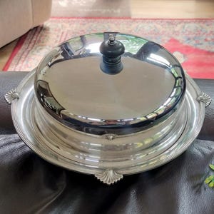 May include: A round silver-toned metal serving dish with a matching domed lid. The lid features a small, dark, rounded knob handle at the centre. The base tray has a wide, flat rim decorated with four evenly spaced shell-like embossed accents.