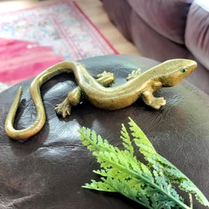 May include: A brass lizard figurine with a curved tail and a small green stone set into the eye. The metallic surface has a polished finish with visible texture along the body, legs, and feet, capturing the form of a reptile in a resting position.