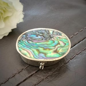 May include: An oval-shaped metal pill box featuring an inlaid abalone shell lid. The shell displays iridescent swirls of green, blue, and purple hues. The box has a polished silver-tone metal base with a small hinged latch on the front side.