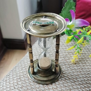 May include: A vintage brass hourglass with a clear glass chamber containing sand. The hourglass features a decorative top with the word "NEWQUAY" in green. The frame is supported by four ornate pillars. The hourglass is a classic timekeeping device.