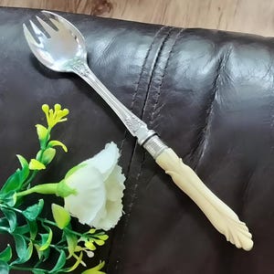 May include: A silver-toned salad serving fork with a polished metal bowl featuring three tines and a decorative embossed neck. The handle is made of a smooth, cream-coloured material with a sculpted, leaf-like design at the base and a ribbed collar.