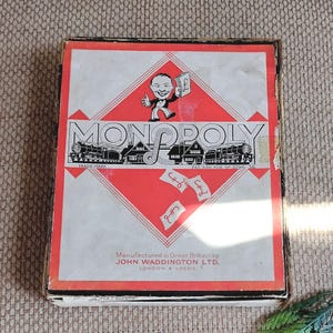 May include: A vintage Monopoly board game box featuring a red diamond design on a white background. The box displays a cartoon character holding a pound symbol, steam trains, houses, and the text Monopoly, Manufactured in Great Britain by John Waddington Ltd, London & Leeds, and Pat. App. For No. 3796-36.