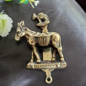 May include: A vintage brass door knocker in the shape of a donkey. The knocker has a donkey figure with a basket and a smaller donkey on top. The word "CLOVELLY" is inscribed on the base. A decorative item.