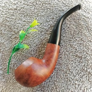 May include: A tobacco pipe with a polished brown wooden bowl and a curved black stem. The word REJECT is stamped into the side of the wooden bowl. A small artificial green stem with yellow flower buds is positioned next to the pipe.