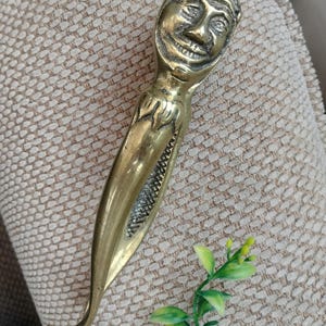 May include: A brass nutcracker shaped like a stylized figure with a smiling face at the top and a textured, tapered body. The metal tool features a hinged design with a pointed base and a smooth, polished finish, showcasing intricate facial details.