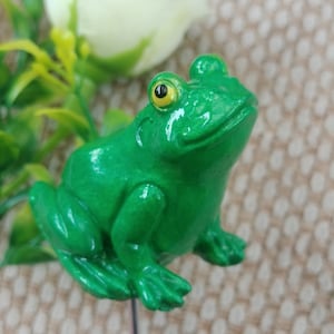 Könnte beinhalten: Eine grüne Froschfigur mit glänzender Oberfläche und einem gelben Auge. Der Frosch sitzt auf einem Stock, der auf dem Bild nicht zu sehen ist.