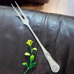 May include: A silver-toned metal cocktail fork featuring two long, slender tines and a decorative handle with an embossed shell motif at the base. The utensil has a polished finish and a tapered stem, showcasing intricate scrollwork near the handle.
