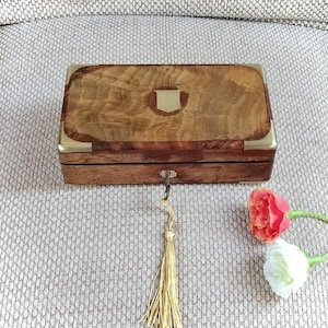 May include: A rectangular wooden box with a polished brown grain finish, featuring brass corner accents and a central brass shield-shaped inlay on the lid. A key with a long gold-colored tassel hangs from the front keyhole of the box.
