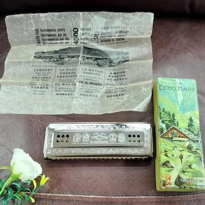 Könnte beinhalten: Eine silberfarbene Hohner Echo Harp Mundharmonika aus Metall mit verzierten Gravuren, begleitet von der originalen rechteckigen Schachtel mit einer Berglandschaftsillustration und einem gefalteten Informationsblatt mit Fabriktext.