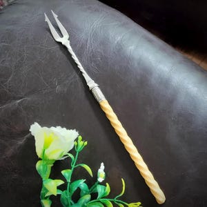 May include: A silver-toned metal carving fork featuring a two-pronged head and a twisted metal neck. The handle is long, cylindrical, and pale yellow with a continuous spiral-twisted texture, connected to the fork by a decorative silver collar.