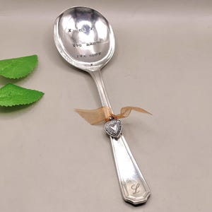 Könnte beinhalten: Ein silberner Löffel mit dem gestempelten Text "X MUM X YOU ARE THE BEST". Ein kleiner Herzanhänger und ein Band sind am Griff befestigt. Der Griff hat den Buchstaben "L" eingraviert.