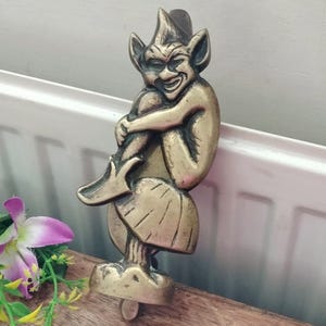 May include: A brass door knocker shaped like a mischievous imp or goblin figure sitting on a mushroom. The figure has pointed ears, a wide grin, and is hugging its knee. The metal has a polished finish with darkened recessed areas for detail.