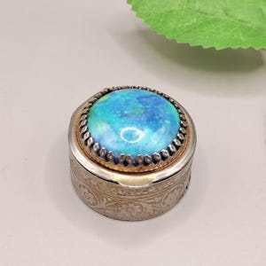 May include: A small, round silver trinket box with a turquoise and blue stone top. The box has a decorative floral pattern around the sides and a gold-colored rim. The stone has a marbled appearance.
