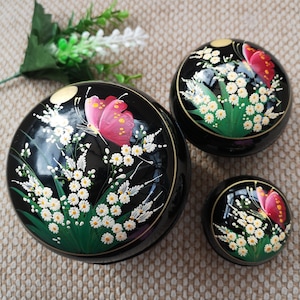 May include: Three black round boxes with lids, each decorated with a white floral design and a pink butterfly. The boxes are nested inside each other, with the largest box at the bottom and the smallest box at the top.