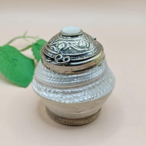 May include: A small, ornate silver-toned trinket box with a textured, pearlescent white base. The lid is detailed with floral patterns and a small, round, white stone. The box is likely used for storing small jewelry items or keepsakes.