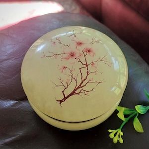 Könnte beinhalten: Eine runde, cremefarbene Dose mit Deckel, die mit einer zarten, handgemalten Illustration eines blühenden Zweigs mit hellrosa Blüten und dunkelbraunen Stielen verziert ist. Die Oberfläche des Behälters hat ein glattes, glänzendes Finish.