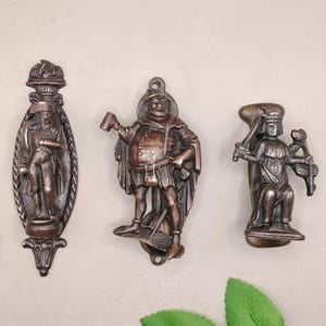 May include: Three antique bronze door knockers. The first features a soldier figure in an oval frame. The second depicts a man holding a mug. The third shows a seated figure with a sword and flower.