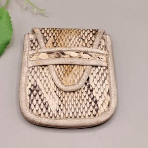 May include: A small, rectangular pouch with a flap closure. The pouch is made of snakeskin in shades of beige, brown, and black. The edges are trimmed with a lighter-coloured leather. The pouch has a unique, textured appearance.