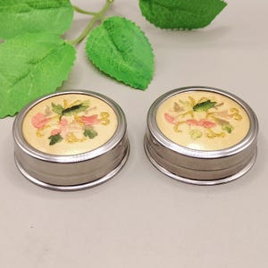 May include: Two small, round, silver-toned containers with embroidered floral designs. The lids feature a cream-colored fabric with pink, green, and yellow floral embroidery. The containers are set against a neutral background with green leaves.