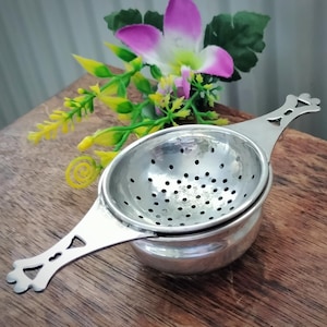 May include: A silver-toned metal tea strainer featuring a perforated bowl and two flat, decorative handles with cut-out details. The strainer is designed with a rounded base and rests on a wooden surface, showcasing a polished finish and functional design.