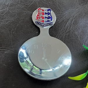 May include: A silver-toned metal souvenir spoon featuring a round bowl engraved with a clock face showing numbers one through twelve and two hands. The handle is topped with a shield-shaped emblem displaying red and blue sections with the text G.T. YARMOUTH.