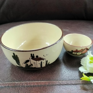 May include: A cream-coloured ceramic bowl and a smaller matching cup, both featuring a hand-painted brown landscape design depicting a small house and trees. The rims of both pieces are finished with a thin, dark brown painted line.