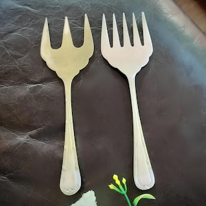 May include: Two silver-toned metal serving forks with long, slender handles featuring vertical line detailing. The left fork has three wide tines, while the right fork has four tines. Both utensils have a smooth, polished finish and a rounded base.