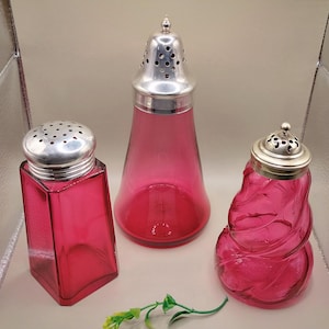 May include: Three antique glass shakers in varying shapes and sizes. The shakers are a vibrant pink colour with silver-coloured metal lids. One is square, one is conical, and the third has a twisted design. A sprig of greenery is at the bottom.