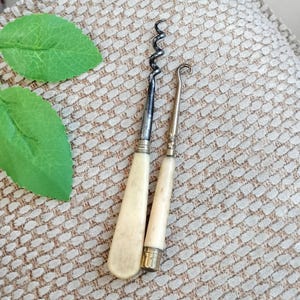 May include: Two antique wine tools: a corkscrew and a hook-shaped tool. Both have off-white handles and metal components. The corkscrew has a spiral design, while the hook tool has a curved end. The tools are displayed on a patterned surface.
