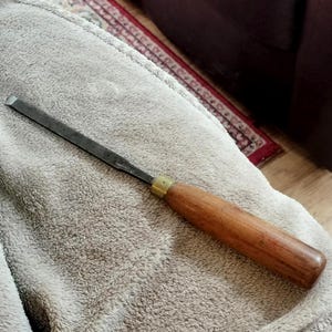 May include: A wood chisel featuring a long, narrow, dark metal blade with a beveled edge and a smooth, cylindrical wooden handle. The handle is connected to the metal shaft by a small, circular brass ferrule, providing a secure grip for woodworking.