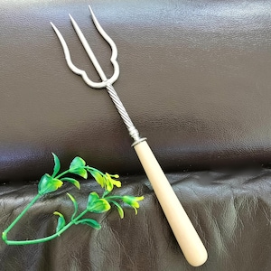 May include: A silver serving fork with three prongs and a decorative handle. The handle is made of light-coloured wood. The fork has a twisted metal section connecting the prongs to the handle. The fork is likely for serving food.