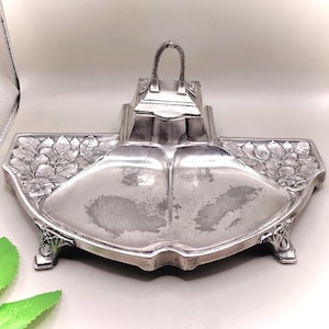 May include: A silver-toned Art Nouveau inkwell and pen tray. The tray has a central inkwell with a handle, flanked by two curved pen rests. Floral and leaf motifs decorate the edges and feet of the tray. The piece is made of metal.