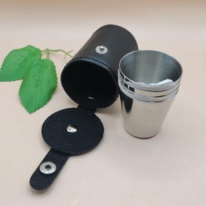 May include: A black leatherette carrying case with a snap closure, open to reveal a set of three stainless steel shot glasses. The case is cylindrical and the shot glasses are stacked. The background is a light beige.