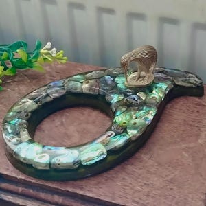 May include: A decorative, dark green, paisley-shaped object inlaid with iridescent abalone shells. A small, gold-colored kiwi bird figurine sits atop the paisley. The object is displayed on a wooden surface.