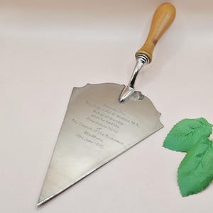 May include: A silver trowel with a wooden handle. The trowel has a decorative, scalloped edge and text that reads "Presented to The St. Rev. R.C.C. Watson M.A., Bishop of Burnley..." The date "21st June 1975" is also visible.