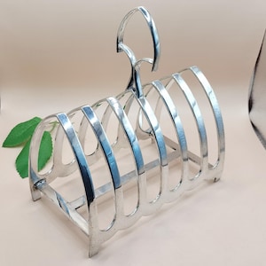 May include: A silver-colored toast rack with a handle. The rack has a rectangular base and curved dividers to hold slices of toast. The handle is arched and attached to the top of the rack. The design is simple and elegant.